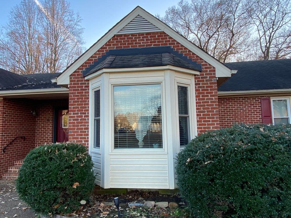 Brick house exterior with large bay window and landscaped shrubs. Clear blue sky in background.
