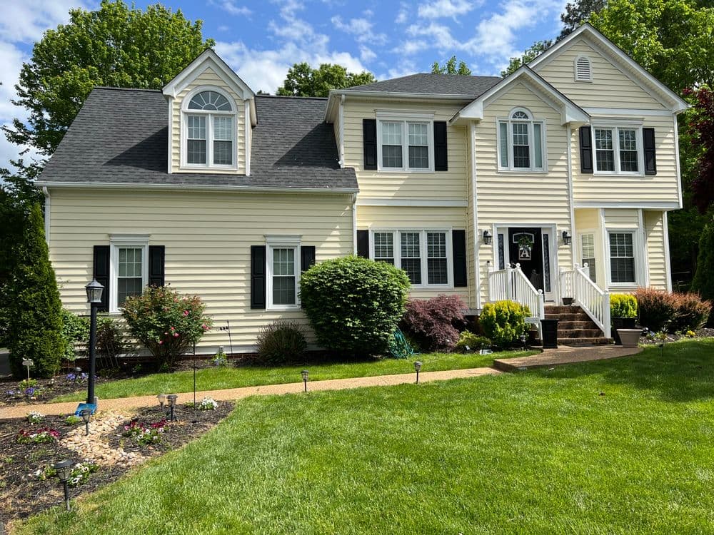 Two-story beige house with black shutters, front steps, and landscaped yard under a blue sky.