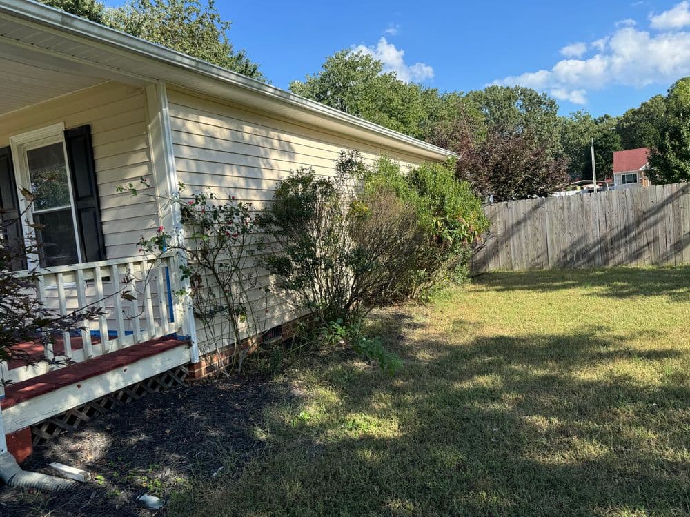 Side view of a beige house with a porch, greenery, and a wooden fence under a clear blue sky.