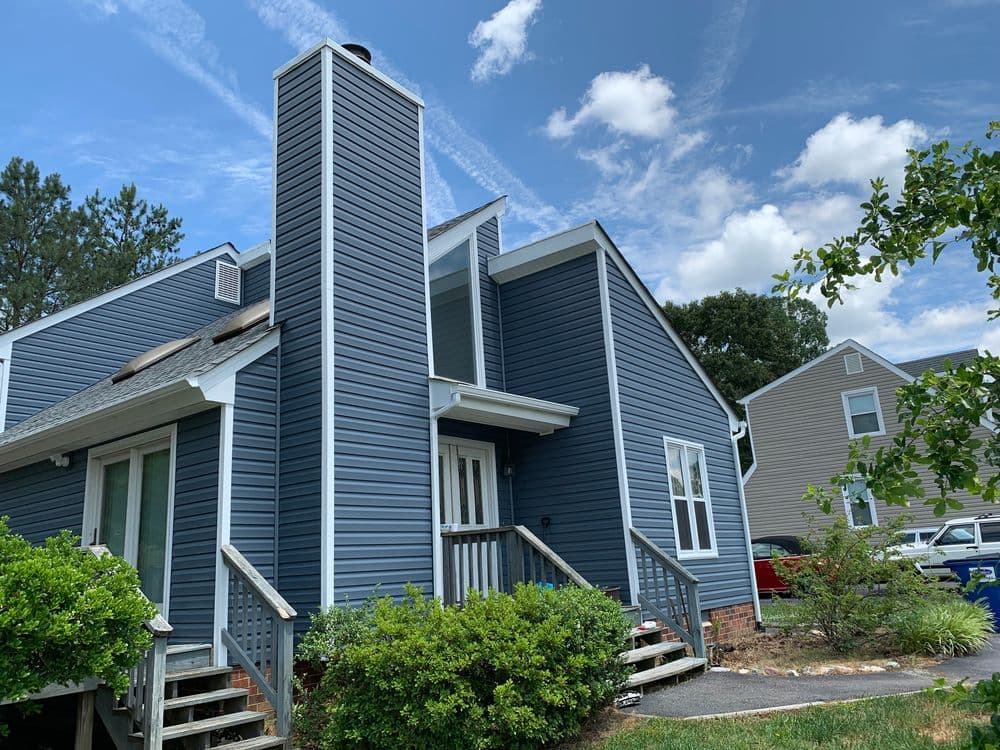 Modern blue house with a tall chimney, surrounded by greenery under a clear sky.