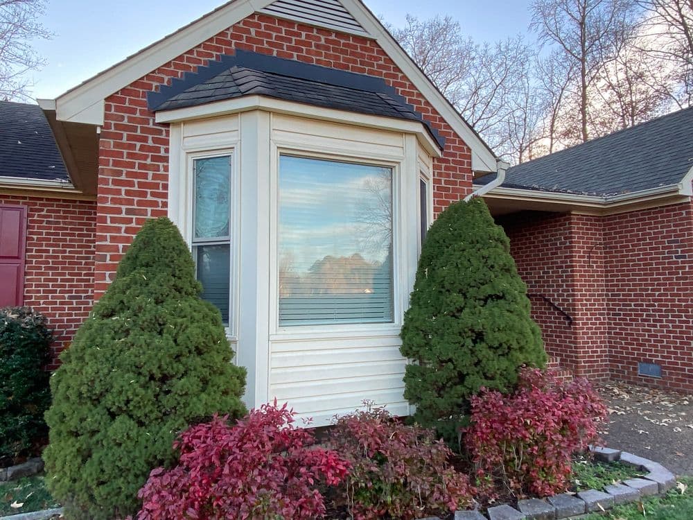 Front view of a brick house featuring a large bay window, shrubs, and colorful flowers.