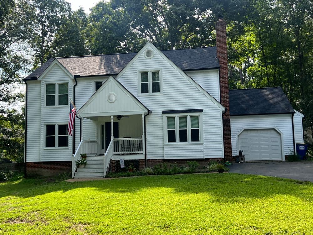 White two-story house with porch, American flag, and landscaped yard surrounded by trees.