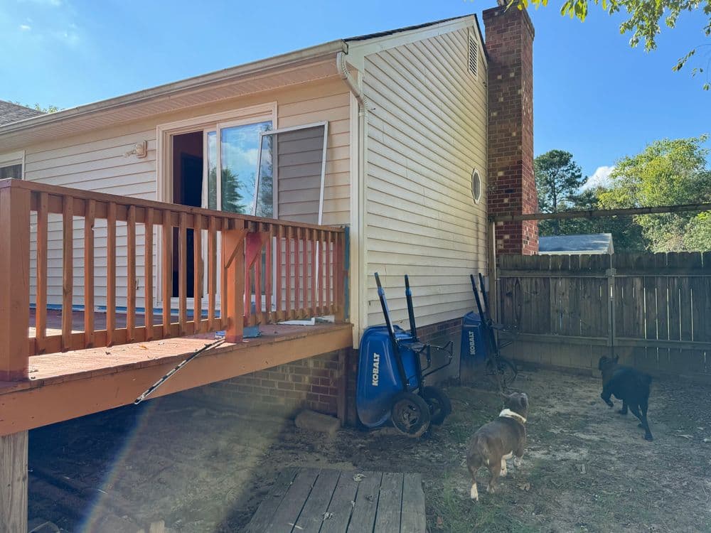 House exterior with a deck, dogs in yard, and garden tools against a wooden fence.